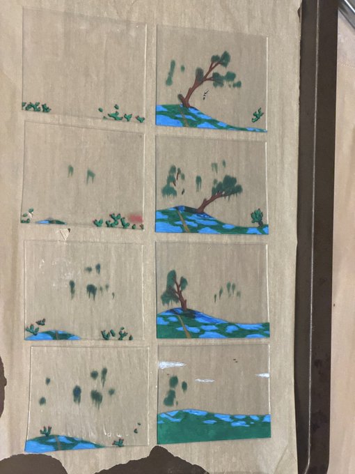 A sheet of glass panels painted with landscape scenes featuring trees and greenery in green and blue hues, mounted on wax paper and positioned next to a metal baking tray.