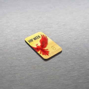A gold-colored "#XRP phoenix badge" with "XRP WEEK" engraved alongside a red phoenix design rests on a brushed metal surface, visually representing the limited-edition physical rewards available through Sl8 Social's XRP Week promotion for participants who bridge or stake qualifying amounts.