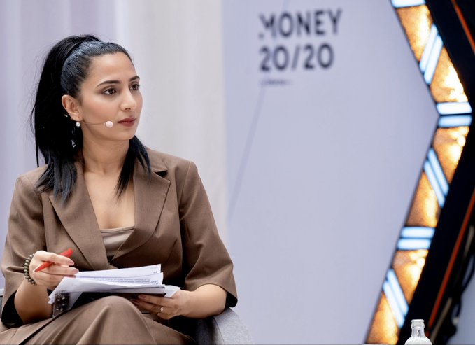 The image shows a woman sitting down with her hands on a piece of paper while wearing a brown blazer. She appears to be engaged in a discussion or presentation, possibly related to the future of private and public blockchains. The background features a white wall with a logo for Money20/20 Europe, which suggests that this event is taking place at a conference or gathering focused on blockchain technology.