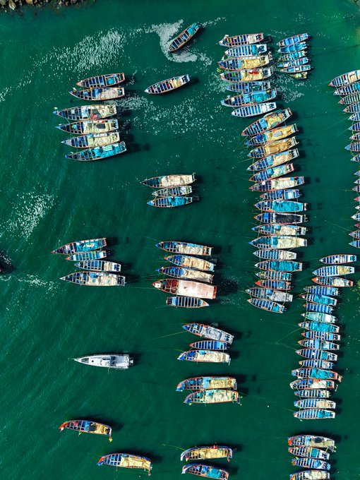 An aerial view of numerous colorful wooden boats clustered together in turquoise coastal waters.