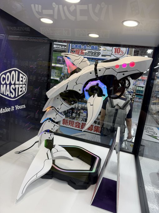 A custom Cooler Master display features an elaborate white and black robotic dog sculpture with glowing pink and RGB accents, displayed inside a glass case at a Japanese electronics store. The backdrop includes Cooler Master branding with the tagline "Make it Yours," along with Japanese promotional signs advertising sales and new memberships.