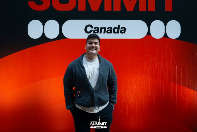 A smiling man wearing a gray cardigan stands before a red backdrop with "Canada" and "Web Summit" visible in white text.