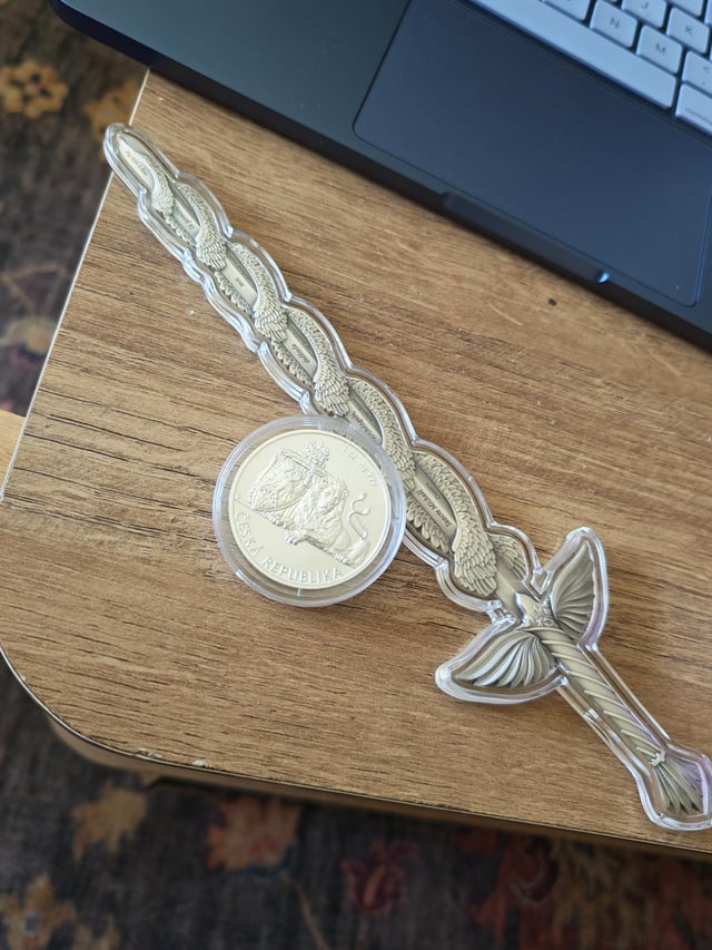 A silver coin labeled "CZECH REPUBLIC" rests on a wooden desk next to an ornate sword design, symbolizing a "buying the dip" moment in the crypto market. The pairing of the coin's detailed artwork with the sword imagery visually represents the post's reference to accumulating assets during market downturns.