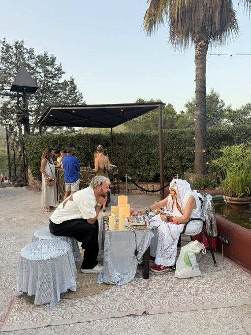 An outdoor scene where a person in white robes sits across a table with candles from a man in a white shirt, with other guests gathered under a shaded canopy in a garden setting.