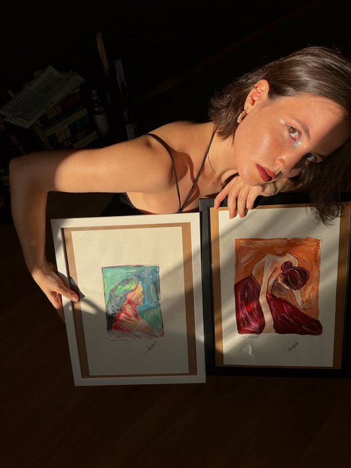 A woman poses in dramatic lighting holding two framed watercolor paintings depicting abstract figures, one labeled "1" and the other "4" with handwritten signatures, likely sharing her weekend art showcase with a cheerful "GM" greeting to friends.