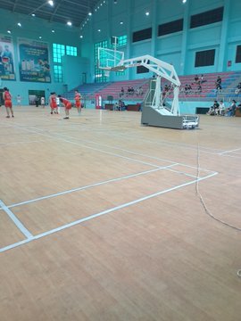 A photograph of an indoor basketball/sports court with wooden flooring, showing players in red and white uniforms on one end and a referee standing on the sidelines.