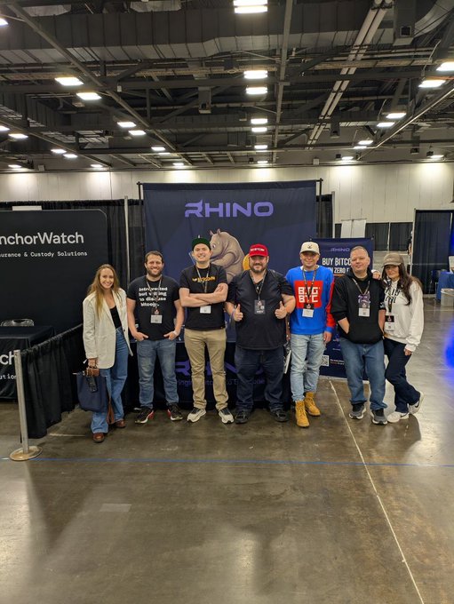 A group of seven people pose for a photo at a convention booth with "RHINO" branding, flanked by "AnchorWatch" and "BUY BITCOIN ZERO" signs.