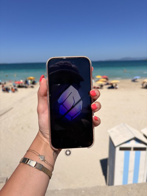 A hand with pink nail polish holds up a smartphone displaying a geometric purple logo against a backdrop of a sunny beach with blue water and a striped beach hut.