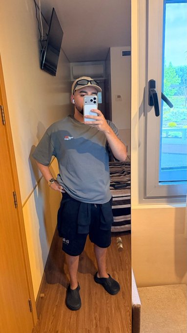 A young man takes a mirror selfie in his room, wearing a gray t-shirt, black shorts, and a beige cap with sunglasses perched on top, while holding up his phone to capture the shot. The "GM" request fits perfectly with this casual, morning-style "Good Morning" post featuring a relaxed home setup with a TV mounted on the wall and a bed visible in the background.