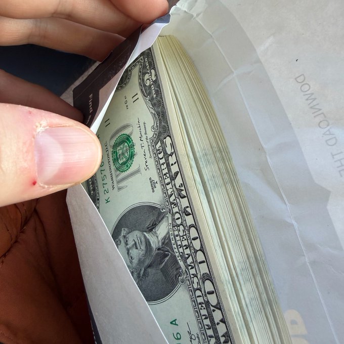 A close-up of a hand flipping through a thick stack of US dollar bills (one and five dollar denominations visible) in what appears to be a delivery envelope marked 'THE DA UNITED'.