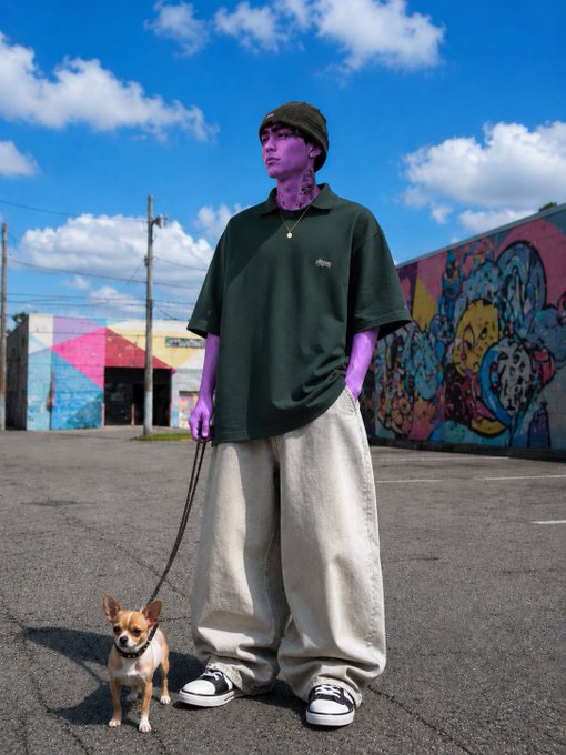 A young man with purple-tinted skin wearing an oversized dark green polo, baggy beige pants, and a beanie, standing with a small chihuahua on a leash in front of colorful street art.