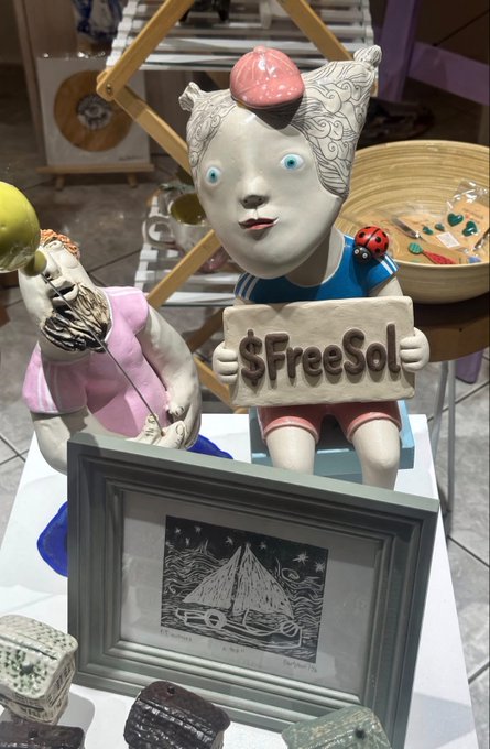 A ceramic sculpture depicts a stylized figure with a pink heart on its head holding a '$FreeSol' sign, accompanied by a framed black-and-white print of a sailboat labeled 'Singer.'