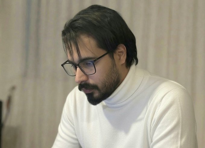 A man with dark hair, a beard, and glasses wearing a white turtleneck sweater, looking down at something off-camera.