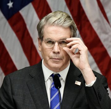 The image shows a man in a dark suit, blue striped tie, and glasses adjusting his glasses with his right hand while speaking, set against an American flag backdrop. He appears to be Treasury Secretary Scott Bessent, whose department's dispute with the Commerce Department is reportedly stalling the implementation of the U.S. Strategic Bitcoin Reserve ordered by President Trump.