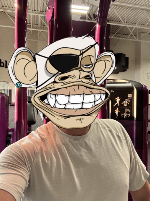 A selfie of a person in a gym with purple equipment, their face obscured by a large cartoon monkey sticker wearing sunglasses and a white cap.