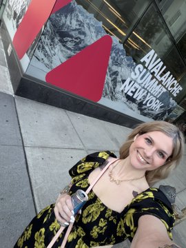 A selfie of a smiling woman in a black and gold patterned top standing outside in front of an Avalanche Summit New York advertisement banner.