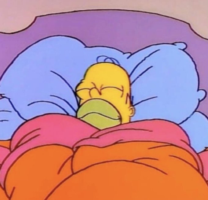Homer Simpson from The Simpsons lies in bed with his eyes closed, appearing sick with green coloring around his mouth, covered by a pink and orange comforter with blue pillows.