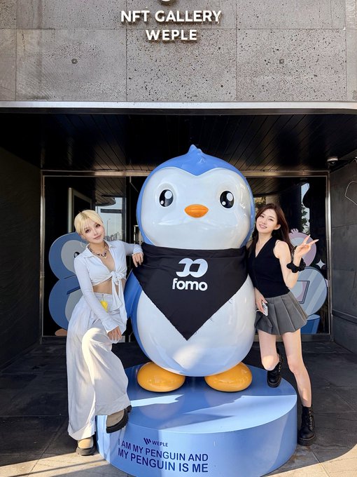 Two women posing on either side of a large penguin mascot statue wearing a 'fomo' bandana, outside the 'NFT GALLERY WEPLE' with text 'I AM MY PENGUIN AND MY PENGUIN IS ME'.