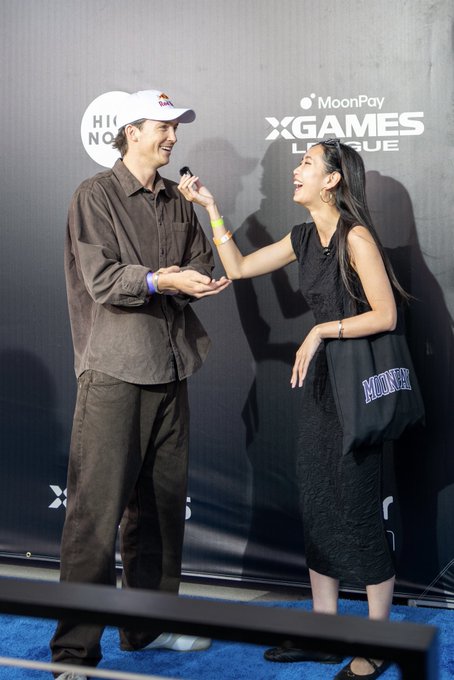 Two people are shown at an event with 'MoonPay' and 'X GAMES LEAGUE' branding in the background; a woman in a black dress interviews a man wearing a brown shirt and a white cap.