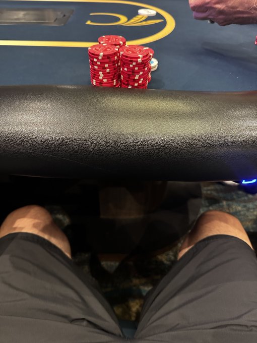 A first-person view of someone sitting at a poker table, looking down past their shorts at stacks of red poker chips and a hand dealing cards.