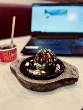 A warm brownie sizzler topped with ice cream and chocolate drizzle sits on a wooden board beside an open laptop, while a small dish to the left displays "Sold Out." The image perfectly captures the post's celebratory moment of enjoying a favorite $3 treat after finishing exams, with the laptop in the background reflecting the trading session mentioned.