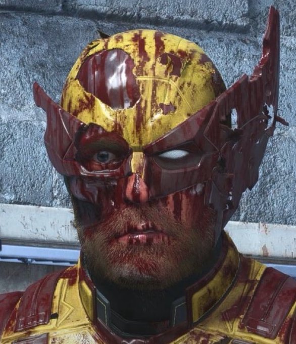 Close-up of a damaged, blood-spattered yellow and red superhero mask with a cracked visor exposing one wide blue eye, with the wearer's bearded face visible underneath against a concrete wall.