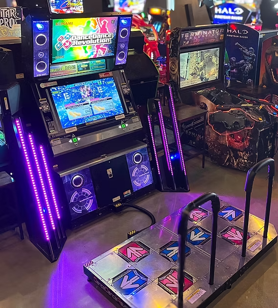ers of a video game arcade, featuring a black and purple console with a large screen and multiple speakers, accompanied by a blue and white platform for motion-based games.