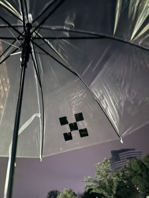 A translucent clear umbrella photographed from below at night, displaying a black pixelated pattern on one of its panels, with trees and a building visible in the background.