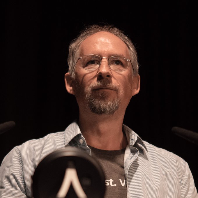 A man with a gray goatee and glasses, identified as Adam Back, looks off to the side during an event, with the partial text "st. V" visible on his gray t-shirt beneath an open light blue shirt. The image relates to the post about his Bitcoin Standard Treasury Company scrapping its 2025 SPAC merger terms with Cantor Equity Partners to renegotiate.