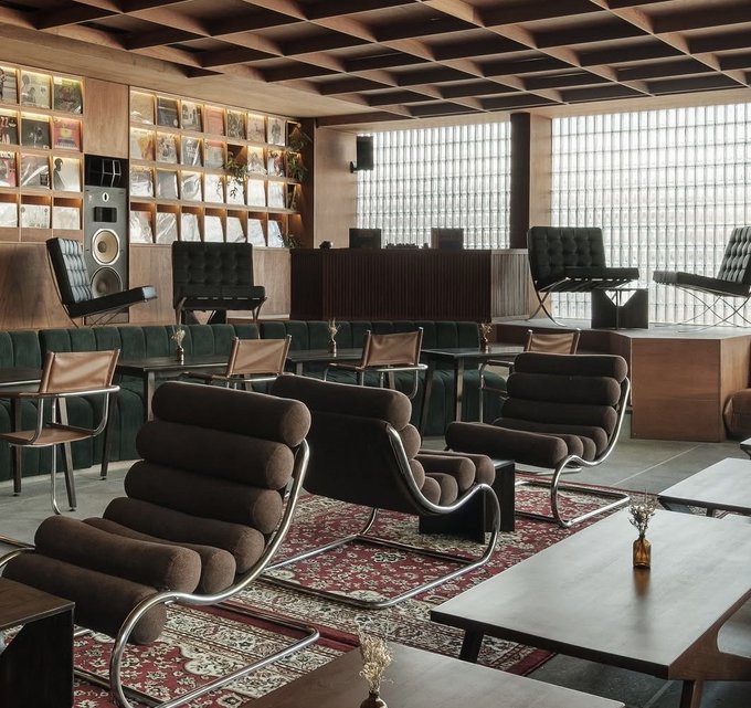 A stylish lounge or café interior featuring brown tubular leather chairs, wooden tables, a Persian-style rug, and a record collection displayed on wall shelves.