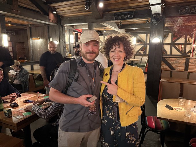 Two people pose for a photo at what appears to be a convention, with the person on the right wearing a yellow jacket and the person on the left wearing a gray t-shirt and cap.