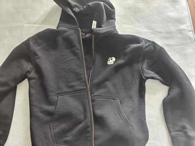 A black zip-up hoodie laid flat on a light-colored surface, featuring a small skull logo on the chest and visible brand tags inside the hood.