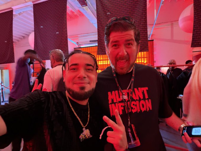 Two men pose together at what appears to be an event; one wears a black 'MUTANT INFUSION' t-shirt and makes a peace sign while the other flashes a rock hand gesture.