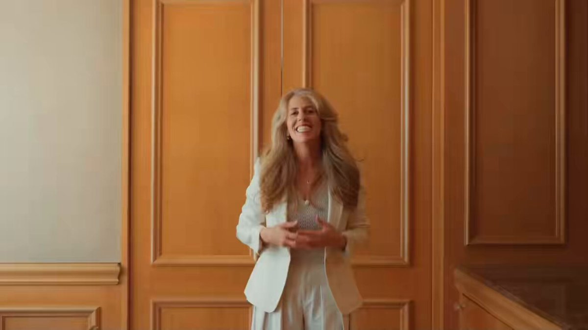 The image features a woman wearing a white blazer, standing in front of wooden doors with her arms raised high above her head. She appears to be smiling and holding something in her hands. The background shows that she is inside an office or a room with wooden walls. There are also some words written on the wall, which seem to be related to the post about the woman's upcoming appearance at a Star Wars convention.