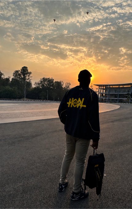 A person stands silhouetted against a vibrant sunset, wearing a black hoodie featuring "HOOL" in bold yellow graffiti-style lettering and holding a black bag while facing away from the camera. The atmospheric scene, with distant kites in the sky and modern buildings on the horizon, invites viewers to piece together the traveler's story from the striking image alone.