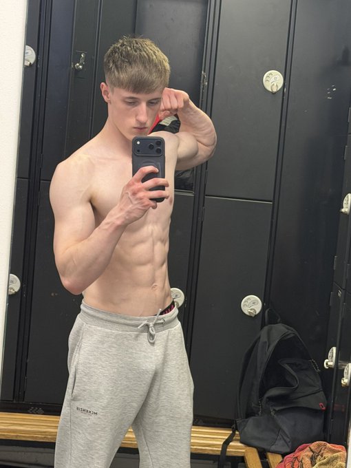 A shirtless, muscular young man takes a mirror selfie in a locker room wearing gray sweatpants, with a black backpack on the bench beside him.