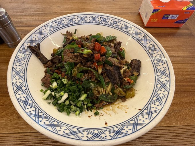 A bowl of stir-fried beef with green onions and red peppers served on a blue and white patterned plate on a wooden table.