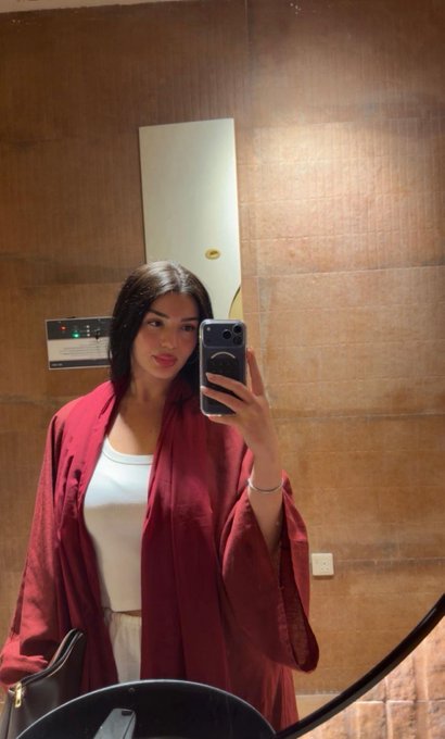 A woman in a red robe over a white top takes a mirror selfie in what appears to be an elevator with wooden panel walls.