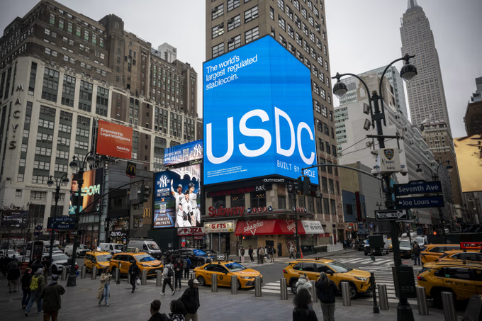 The image shows a busy city street with tall buildings, including one that has a large blue billboard advertising USDC. The street is filled with people walking around, some of whom are carrying backpacks or handbags. There are also several cars parked along the side of the road and moving vehicles driving by. A traffic light can be seen in the background, indicating an urban setting.