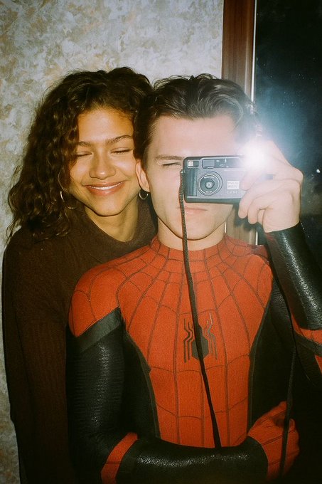 Zendaya and Tom Holland pose together for a mirror selfie, with Holland wearing a Spider-Man suit while Zendaya smiles beside him.