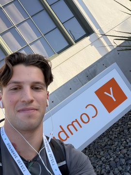 A young man with earphones takes a selfie in front of a sign displaying a 'Y' logo and the partial word 'Comp' outdoors.