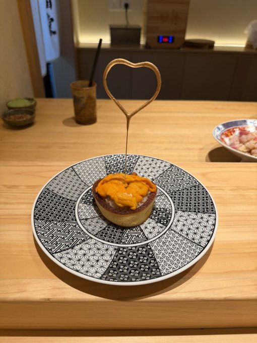 A Japanese dish, appearing to be uni (sea urchin) served over a tart or sushi-style base, presented on a decorative patterned plate with a heart-shaped decorative pick on a wooden counter.