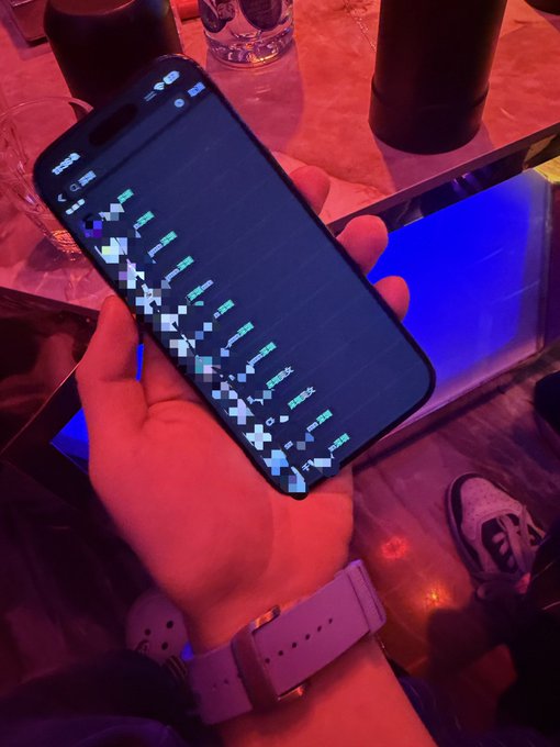 A person holds up a smartphone displaying a grid of green tickers—resembling a stock or crypto trading app—as a flex, with a wristwatch visible and sneakers peeking in from below. The image embodies the caption's boast of "Wealth" and "top-tier charm," showing off the tools and accessories of a self-proclaimed high-roller.