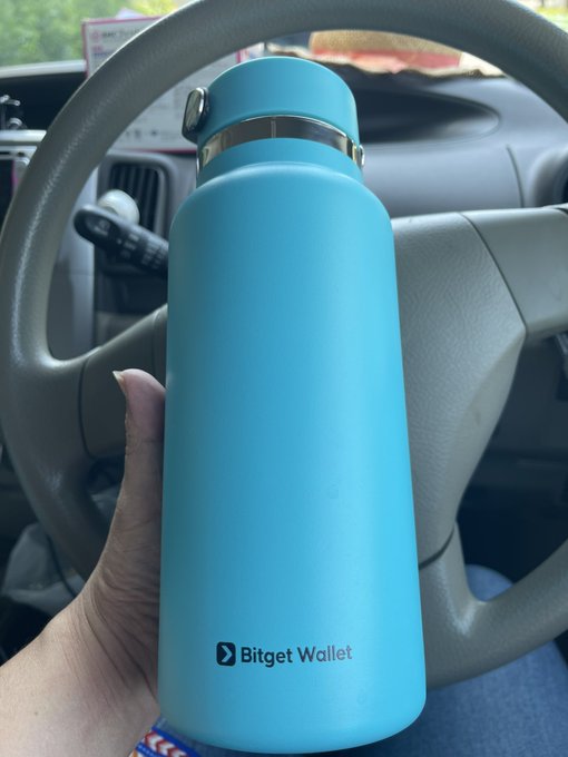 A hand holds a light blue water bottle branded with the "Bitget Wallet" logo, photographed inside a car with the steering wheel visible in the background.
