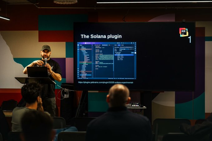 A presenter speaking into a microphone at a tech event, with a screen behind him displaying "The Solana plugin" and a GitHub link for jupiter-terminal-experimental.