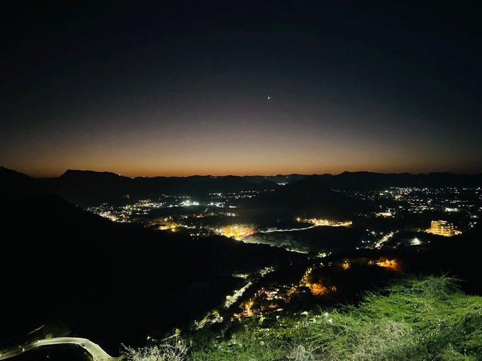 A nighttime view of a city valley with scattered lights, mountains in the distance, and a dark sky.