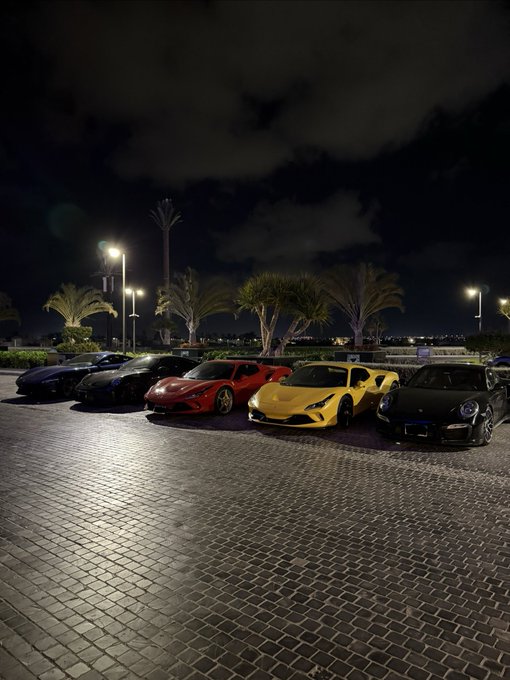 A lineup of luxury sports cars, including Ferraris, a Lamborghini, and a Porsche, parked on a cobblestone lot at night, symbolizing the lavish lifestyle and success that comes with trading on RiseX with the boys.