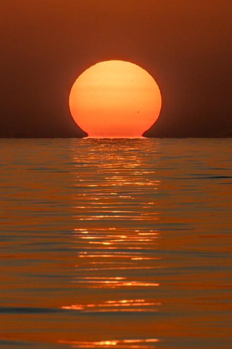 A large orange sun setting over a calm body of water, casting a long, shimmering reflection on the surface.