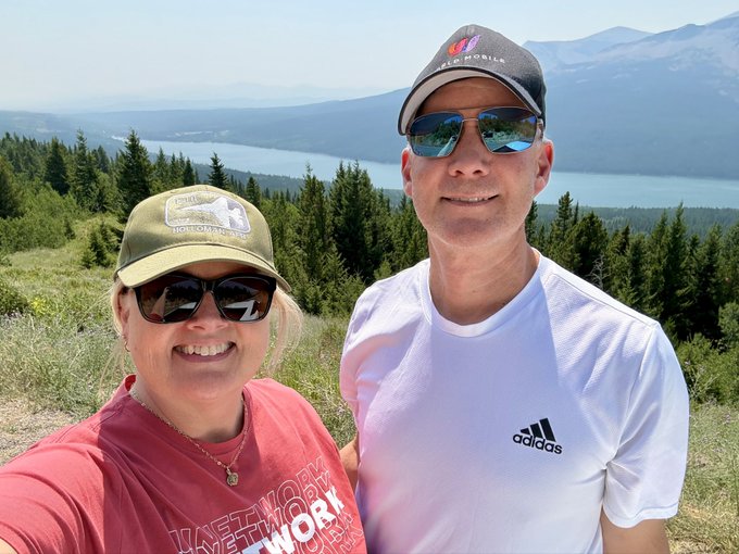 A selfie of a smiling couple wearing caps and sunglasses, with the man in a white Adidas shirt and the woman in a red shirt, posing on a mountain overlook with a scenic lake and forested hills in the background.