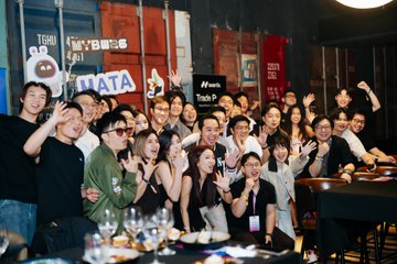 A group of about 15 people smile and raise their hands at a post-event gathering, with visible signage reading "TSG" and "HATA" in the background. The lively photo illustrates the genuine appreciation and warm community support the post describes.